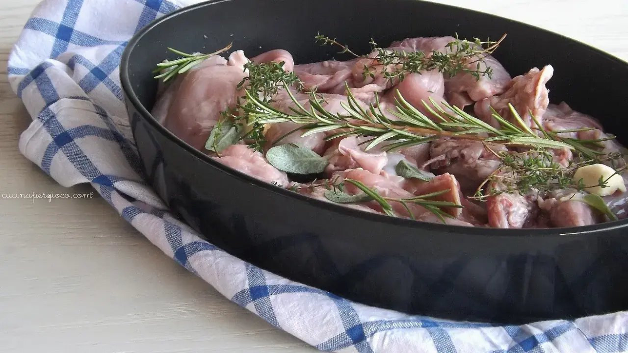 Pezzi di coniglio crudo in una teglia, pronti per essere cucinati al forno con rosmarino, salvia e aglio.