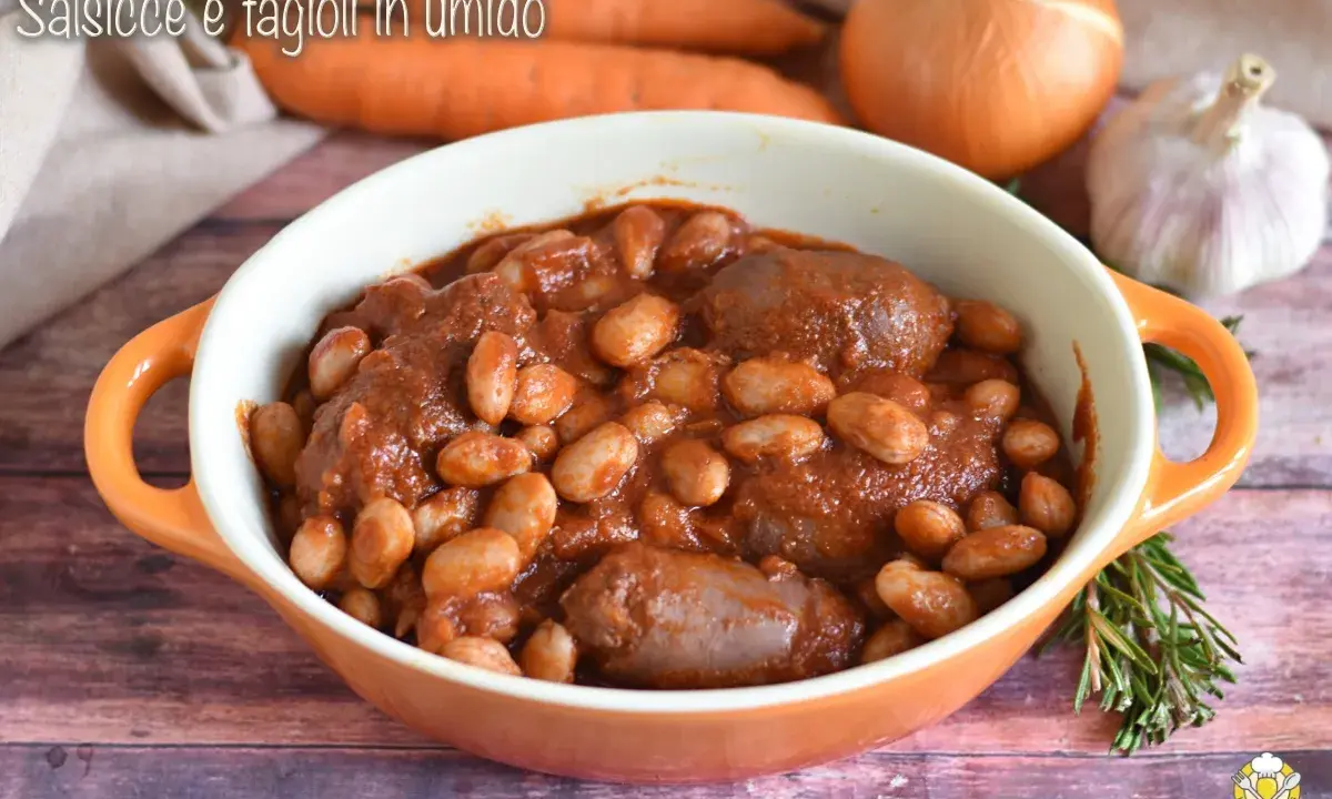 Un saporito piatto di salsiccia e fagioli in umido, servito in una ciotola arancione, con carote, cipolla e aglio sullo sfondo.