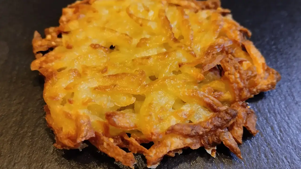 Un croccante rosti di patate dorato, con bordi leggermente bruciacchiati, adagiato su una superficie scura.