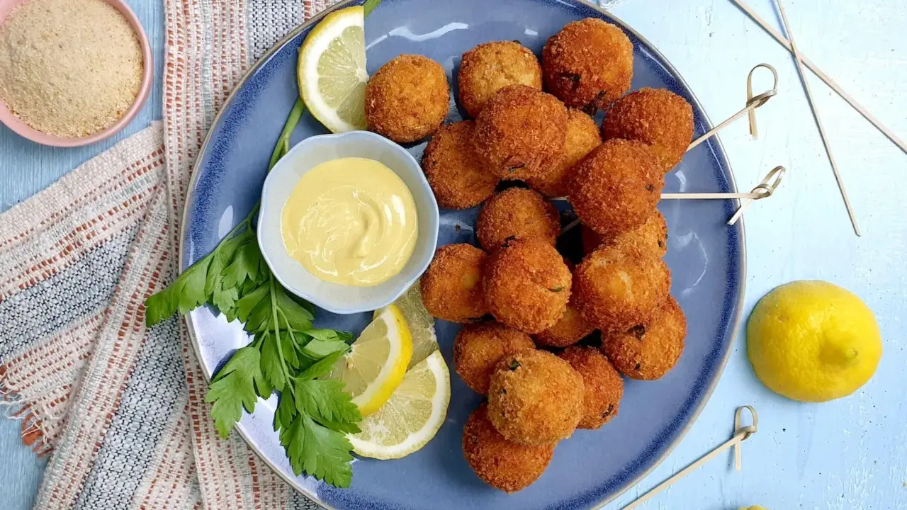 Piatto di polpette di tonno dorate e croccanti, servite con spicchi di limone, prezzemolo e una ciotolina di salsa gialla.