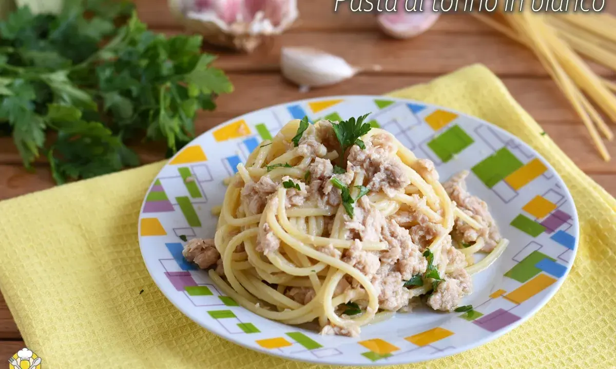 Un piatto di pasta al tonno in bianco, condita con tonno e prezzemolo, su un piatto colorato. Sullo sfondo, aglio e prezzemolo fresco.