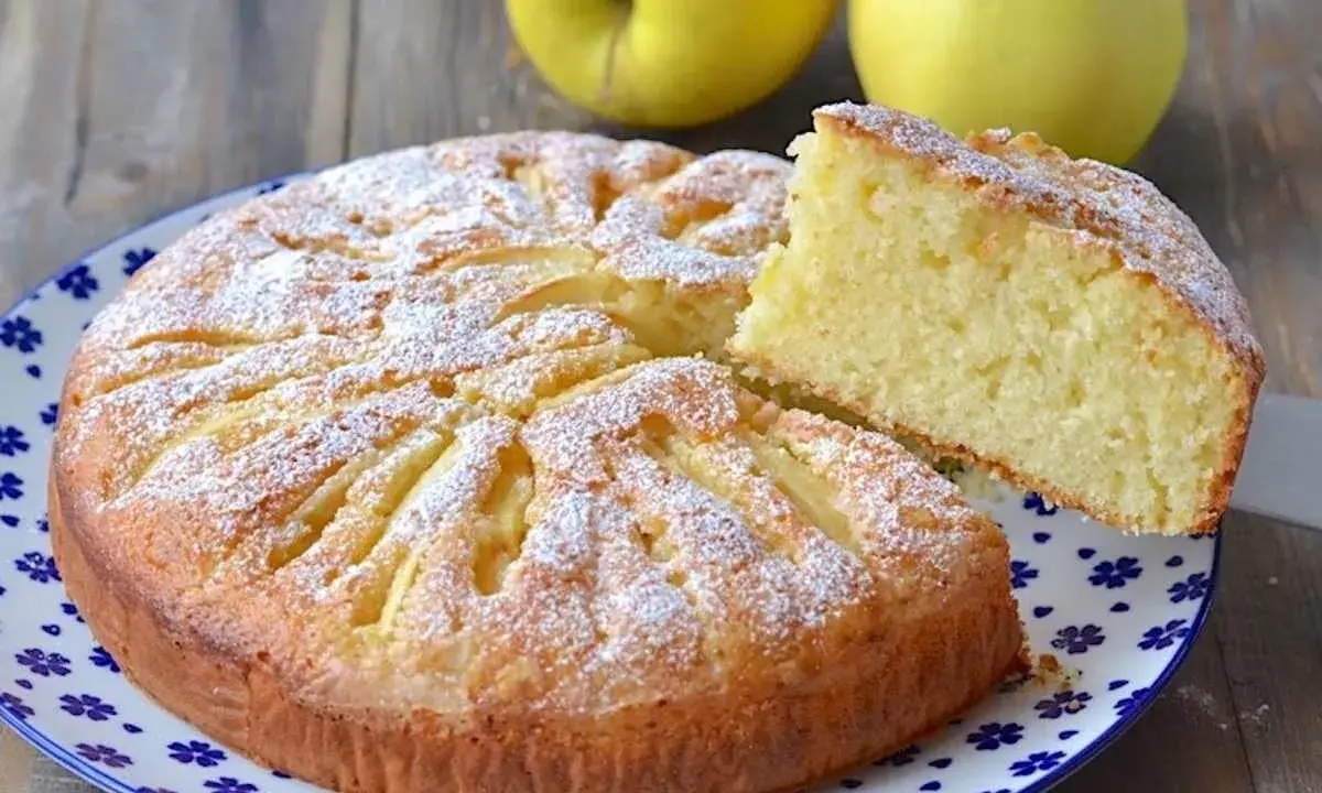 Deliziosa torta di mele con yogurt, cosparsa di zucchero a velo, con una fetta sollevata per mostrare la morbidezza.