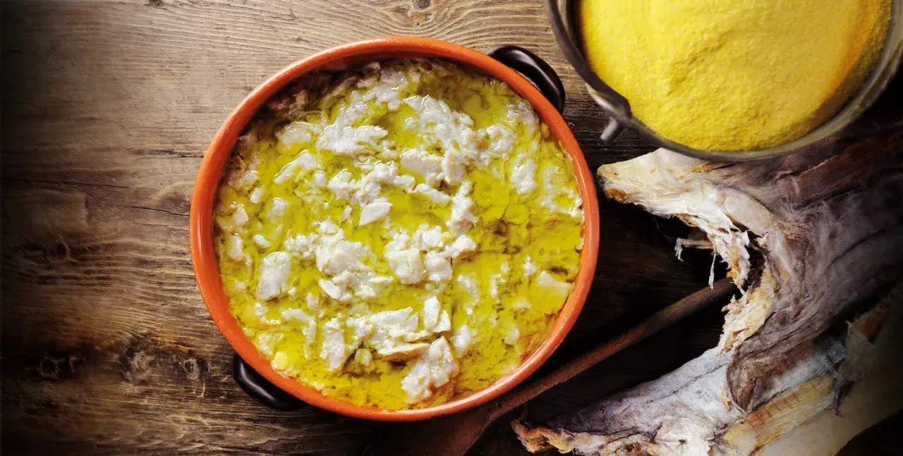 Piatto fumante di baccalà alla vicentina, con polenta gialla e stoccafisso essiccato sullo sfondo.