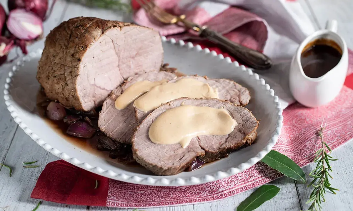 Arrosto di vitello morbido con sughetto benedetta, servito su un piatto bianco con cipolle rosse caramellate e rosmarino.
