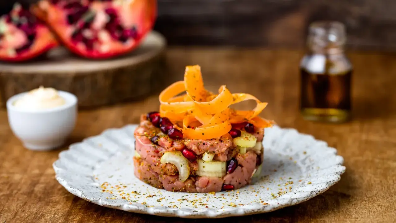 Tartare di manzo con melograno e carote, servita su un piatto rustico. Sullo sfondo, spicchi di melograno e una bottiglietta di condimento.