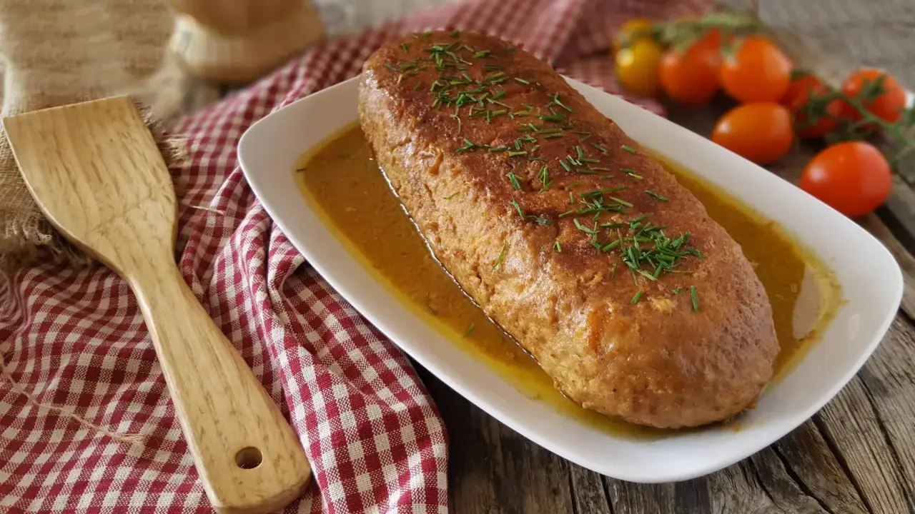 Polpettone in padella dorato e succulento, guarnito con erba cipollina, pronto per essere servito. Accanto, una spatola in legno e pomodorini freschi.