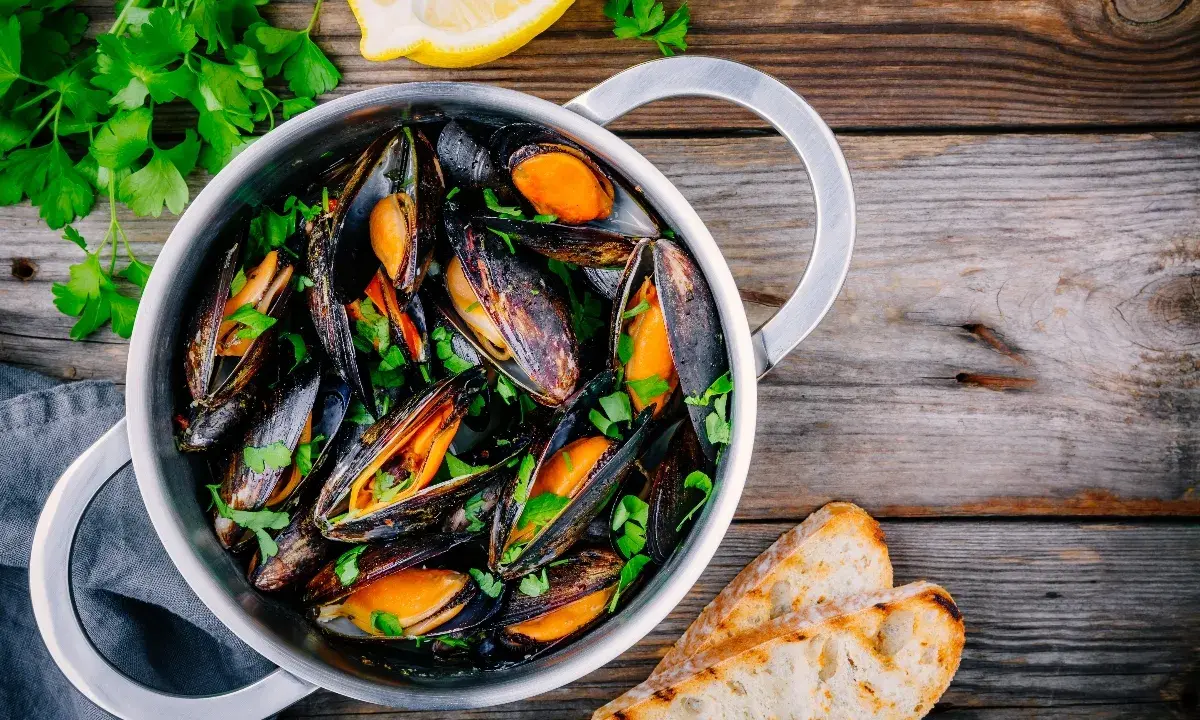 Un pentolino di cozze alla marinara, guarnite con prezzemolo fresco e accompagnate da fette di pane tostato e spicchi di limone.