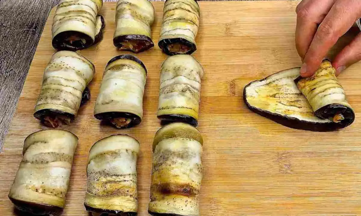 Involtini di melanzane pronti per essere infornati. Una mano sta arrotolando l'ultima fetta di melanzana grigliata.