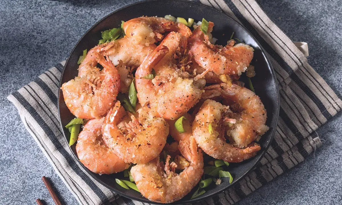 Gamberi sale e pepe croccanti, serviti su un piatto scuro con erba cipollina fresca. Un piatto invitante e saporito.