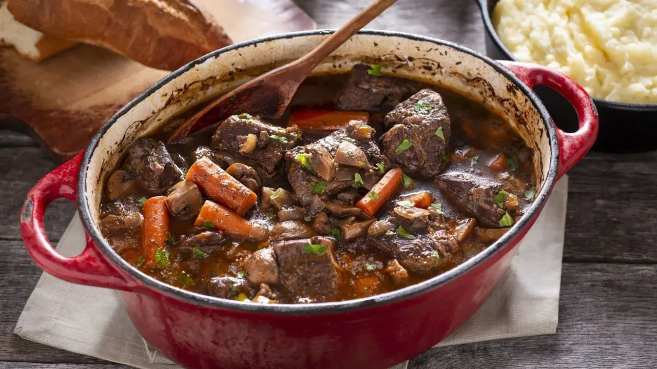Un invitante spezzatino di manzo in un tegame rosso, con carote, funghi e un mestolo di legno. Accanto, pane rustico e purè di patate.