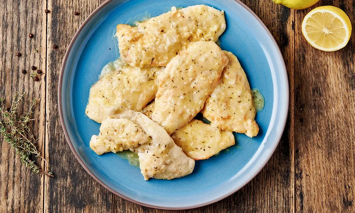 Piatto di pollo al limone su un piatto blu, con limone e rosmarino sullo sfondo.