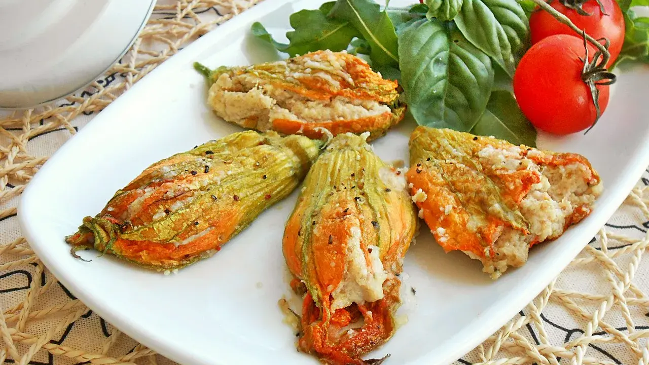 Deliziosi fiori di zucca al forno ripieni, serviti su un piatto bianco con pomodorini e basilico fresco.