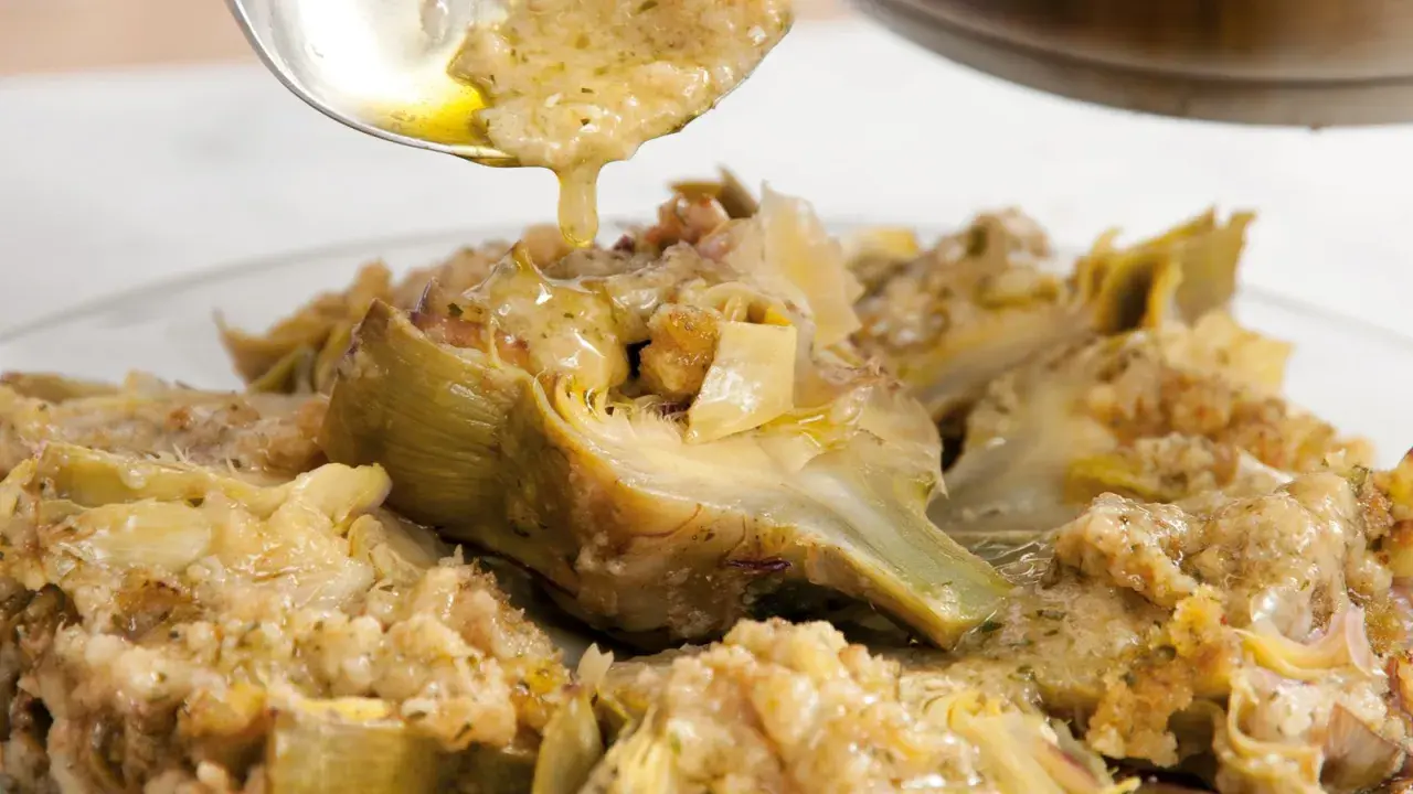 Un cucchiaio versa un sugo dorato su un piatto di carciofi alla romana, conditi con erbe aromatiche e pane croccante.