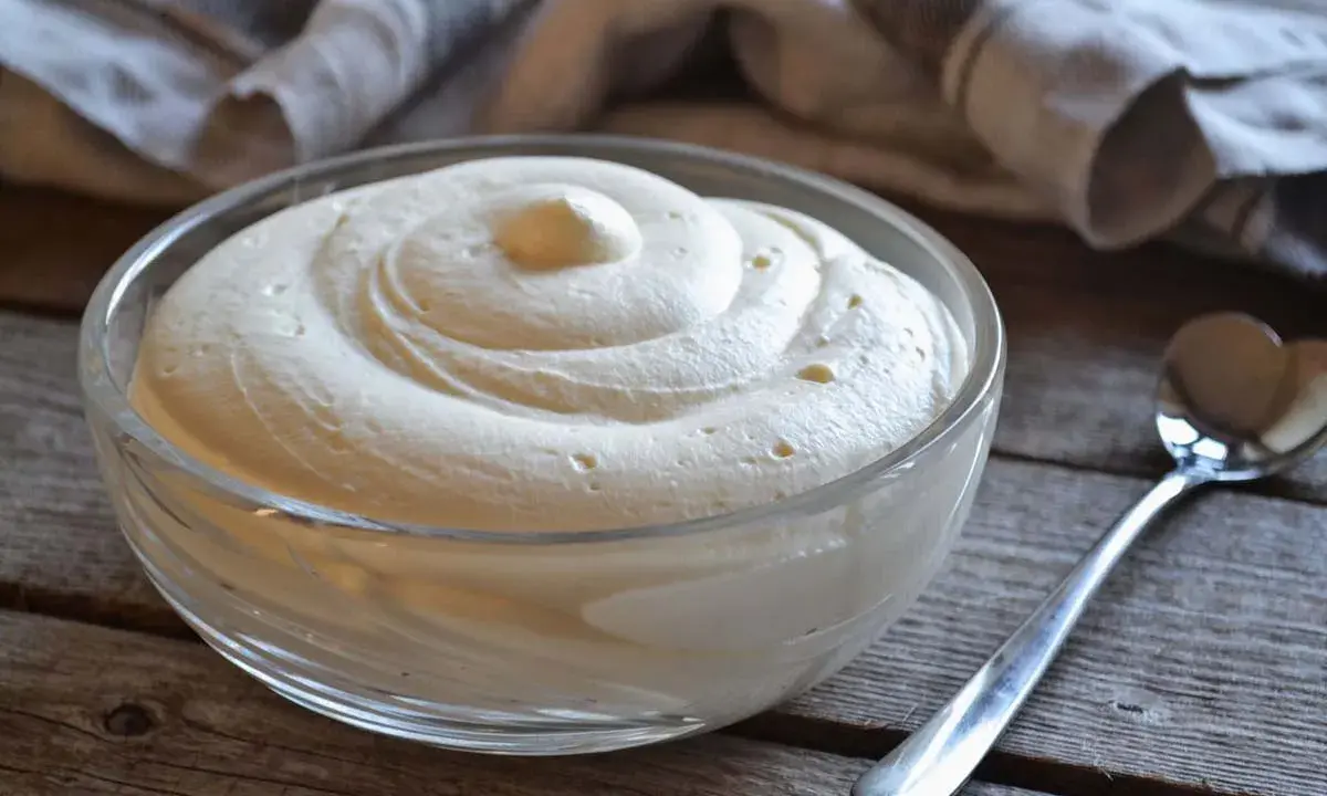 Una ciotola di vetro piena di soffice crema al mascarpone, con una spirale perfetta e una cucchiaio d'argento accanto.