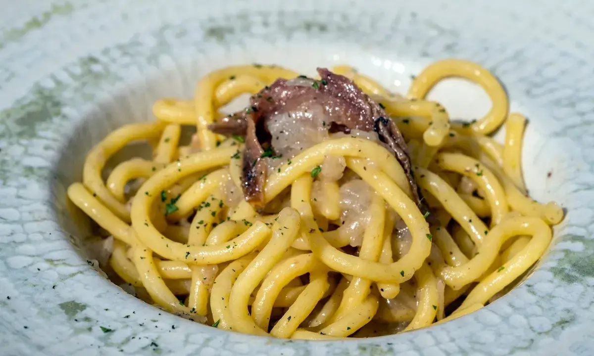 Un piatto di bigoli in salsa, pasta spessa condita con acciughe e cipolla, guarnita con prezzemolo tritato.