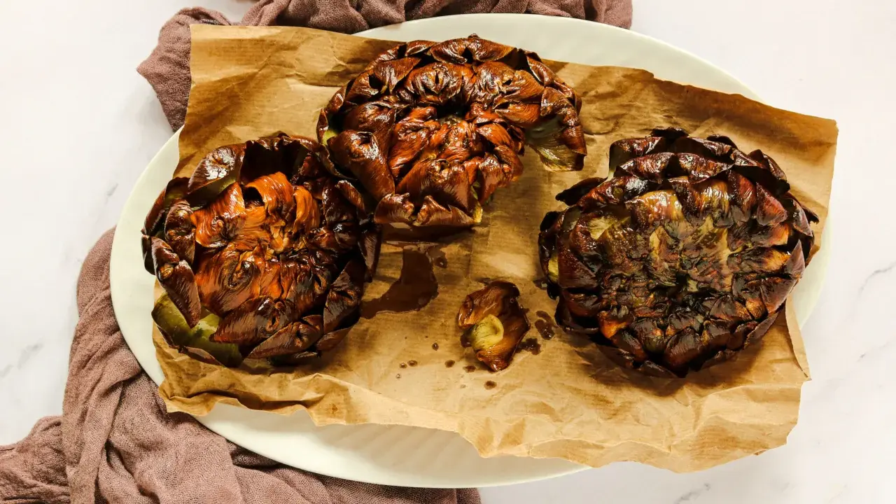 Tre carciofi alla giudia dorati e croccanti, serviti su carta da forno marrone, su un piatto bianco.