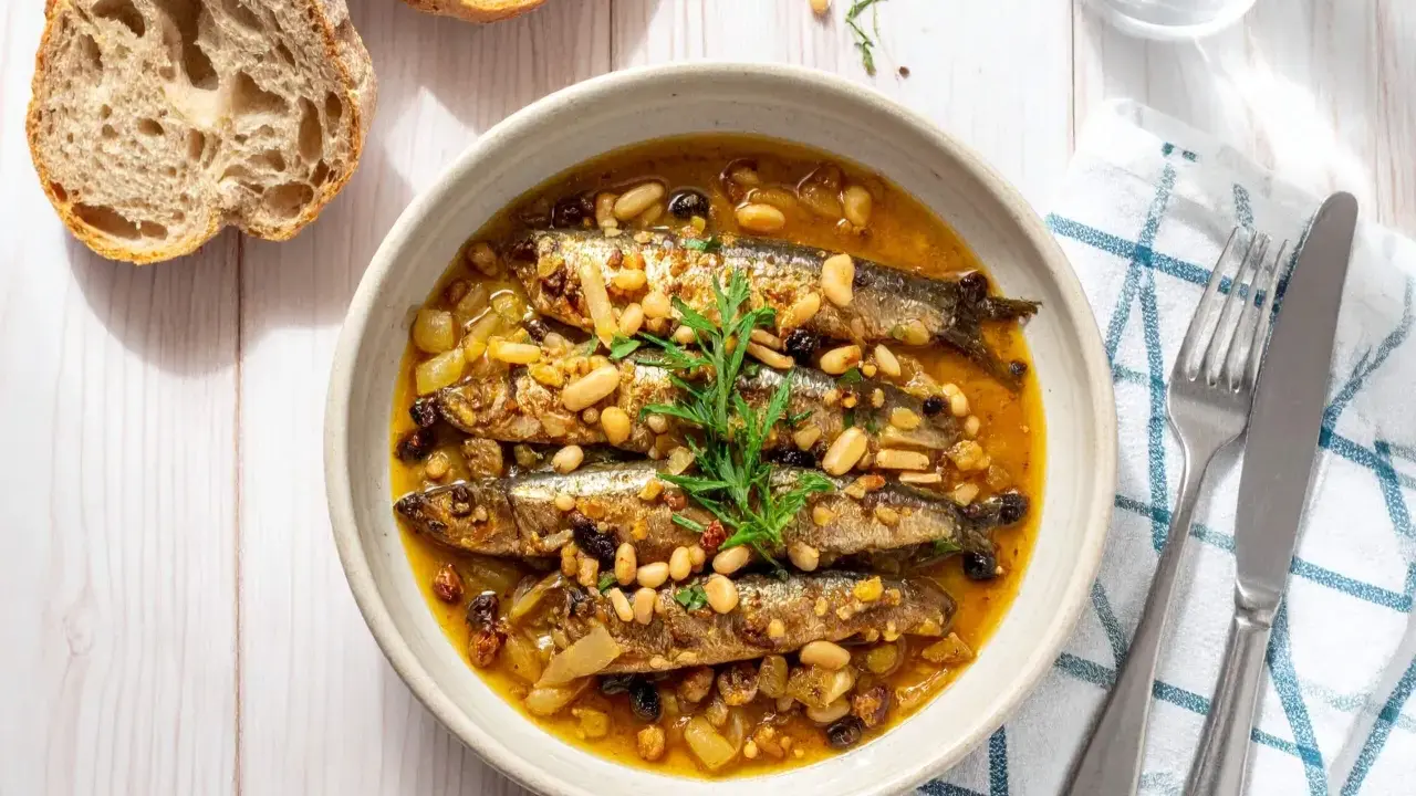 Sarde in saor in una ciotola, con pinoli, uvetta e cipolle. Accanto, pane rustico e posate.