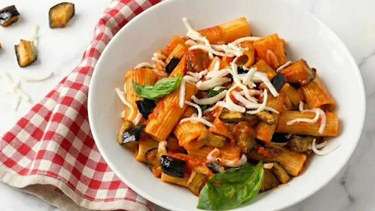 Un piatto di pasta alla norma fumante, con melanzane fritte, pomodoro e ricotta salata grattugiata, guarnito con basilico fresco.