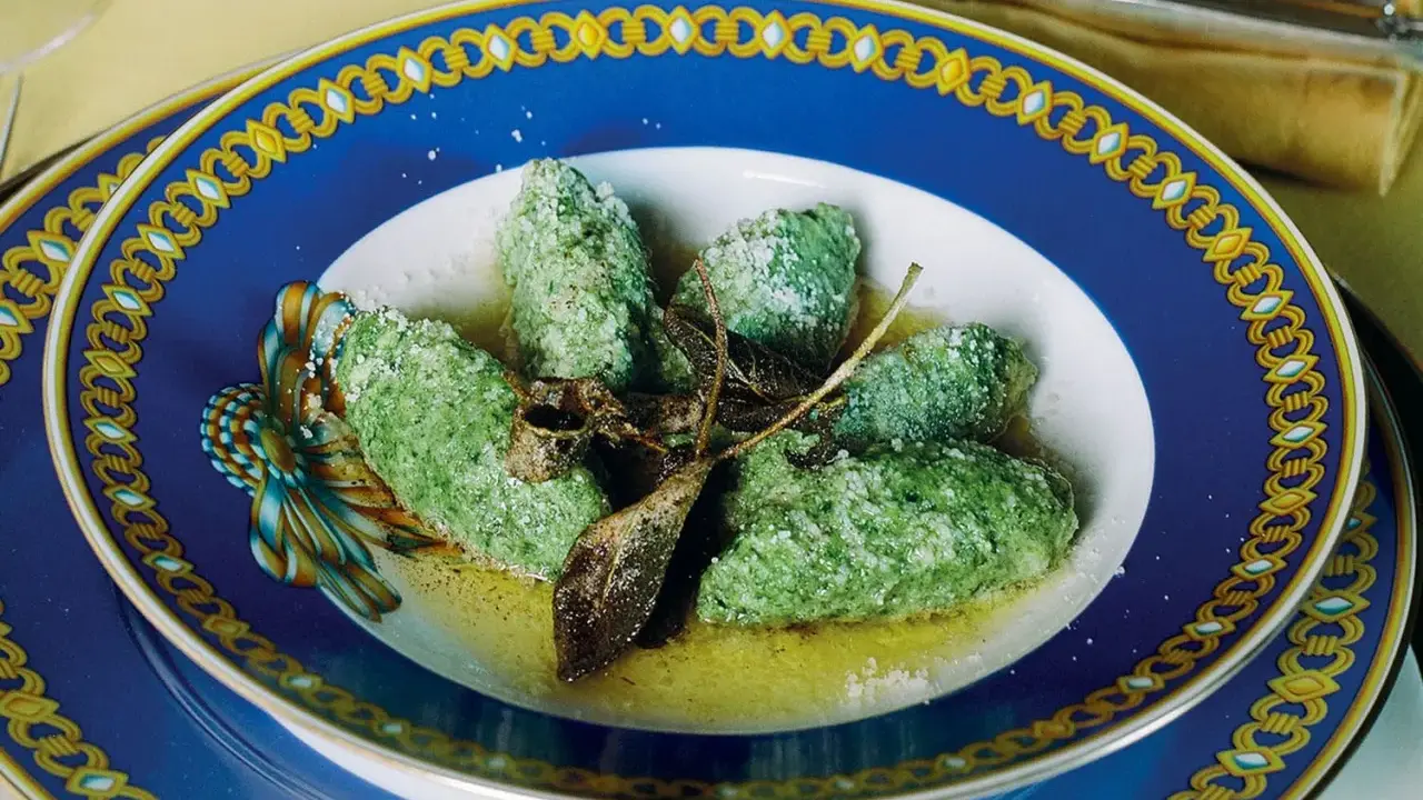 Strangolapreti alla trentina, verdi e saporiti, serviti in un piatto blu decorato con burro fuso e parmigiano.