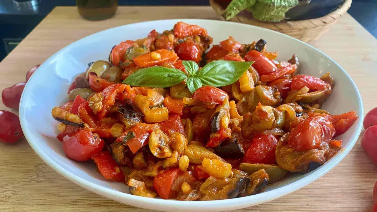 Caponata siciliana ricetta della nonna: melanzane, pomodori, peperoni e basilico in un piatto bianco.
