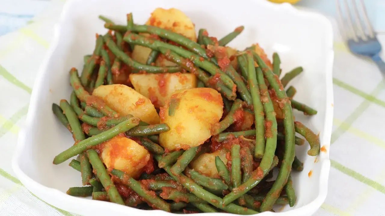 Un piatto di fagiolini in umido con patate, conditi con un sugo rosso saporito, servito in una ciotola bianca.