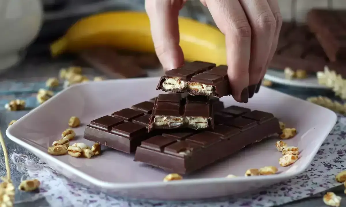A hand breaks a chocolate bar filled with puffed rice and nuts on a pink plate.