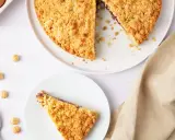 Una fetta di sbriciolata alla Nutella, con la torta intera e ingredienti come nocciole e uova sullo sfondo.