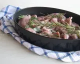 Pezzi di coniglio crudo in una teglia, pronti per essere cucinati al forno con rosmarino, salvia e aglio.