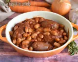Un saporito piatto di salsiccia e fagioli in umido, servito in una ciotola arancione, con carote, cipolla e aglio sullo sfondo.