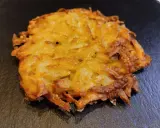 Un croccante rosti di patate dorato, con bordi leggermente bruciacchiati, adagiato su una superficie scura.