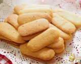Un mucchio di biscotti a forma di lingue di gatto, dorati e friabili, disposti su una tovaglietta di pizzo bianco.