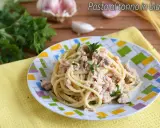 Un piatto di pasta al tonno in bianco, condita con tonno e prezzemolo, su un piatto colorato. Sullo sfondo, aglio e prezzemolo fresco.