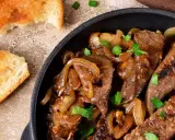 Fegato alla veneziana succulento in padella con cipolle caramellate e prezzemolo fresco, accompagnato da fette di pane tostato.