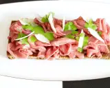 Delicato carpaccio di manzo su crostino integrale, guarnito con crema verde, scaglie di parmigiano e germogli freschi.