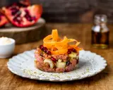 Tartare di manzo con melograno e carote, servita su un piatto rustico. Sullo sfondo, spicchi di melograno e una bottiglietta di condimento.