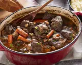 Un invitante spezzatino di manzo in un tegame rosso, con carote, funghi e un mestolo di legno. Accanto, pane rustico e purè di patate.