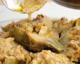 Un cucchiaio versa un sugo dorato su un piatto di carciofi alla romana, conditi con erbe aromatiche e pane croccante.