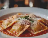 Deliziosi ravioli fatti in casa, conditi con sugo rosso, parmigiano grattugiato e un rametto di erbe aromatiche.