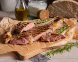 Succulenta costata di manzo alla griglia, tagliata a fette, con rosmarino fresco e sale grosso. Sullo sfondo, pane rustico e olio d'oliva.