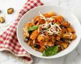 Un piatto di pasta alla norma fumante, con melanzane fritte, pomodoro e ricotta salata grattugiata, guarnito con basilico fresco.