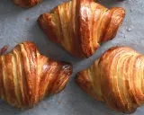 Tre cornetti fatti in casa dorati e sfogliati, appena sfornati, con gocce di condensa sulla carta da forno.