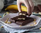 A hand breaks a chocolate bar filled with puffed rice and nuts on a pink plate.