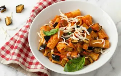 Un piatto di pasta alla norma fumante, con melanzane fritte, pomodoro e ricotta salata grattugiata, guarnito con basilico fresco.