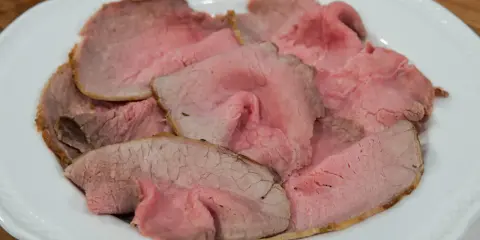 Fette sottili di arrosto di manzo rosa e succulento, servite su un piatto bianco.