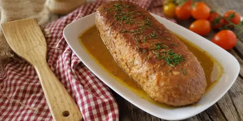 Polpettone in padella dorato e succulento, guarnito con erba cipollina, pronto per essere servito. Accanto, una spatola in legno e pomodorini freschi.