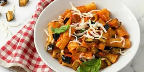 Un piatto di pasta alla norma fumante, con melanzane fritte, pomodoro e ricotta salata grattugiata, guarnito con basilico fresco.