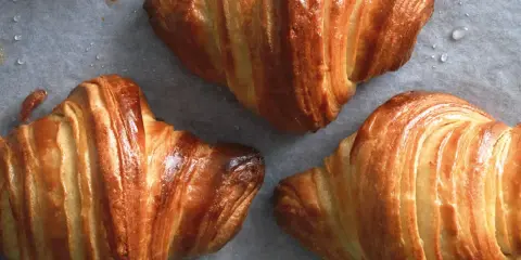Tre cornetti fatti in casa dorati e sfogliati, appena sfornati, con gocce di condensa sulla carta da forno.