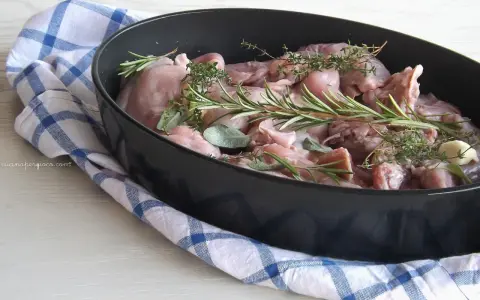Pezzi di coniglio crudo in una teglia, pronti per essere cucinati al forno con rosmarino, salvia e aglio.