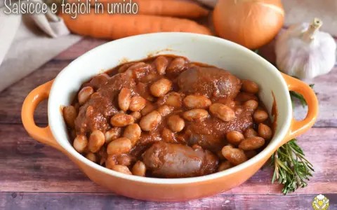 Un saporito piatto di salsiccia e fagioli in umido, servito in una ciotola arancione, con carote, cipolla e aglio sullo sfondo.