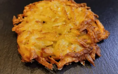 Un croccante rosti di patate dorato, con bordi leggermente bruciacchiati, adagiato su una superficie scura.